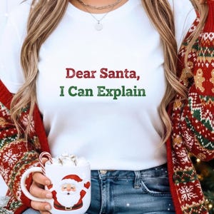 Op de afbeelding: Wit T-shirt met de tekst "Dear Santa, I Can Explain" in rood en groen. De afbeelding toont een persoon die het shirt draagt, een rood vest en een mok met een kerstmanontwerp vasthoudt. De mok bevat slagroom en een zuurstok.