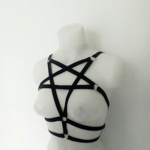 May include: A black elastic body harness featuring a pentagram design across the chest. The harness is constructed from thin fabric straps connected by silver-toned metal rings, forming a geometric star pattern that extends around the torso.