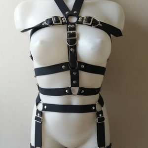 May include: Black body harness with adjustable straps and silver-tone metal hardware. The harness features a halter neck, chest straps, and leg straps, all connected with metal rings and buckles. The design is displayed on a white mannequin.
