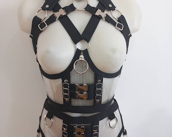 May include: A black body harness featuring elastic straps, silver-toned metal rings, and adjustable buckles. The design includes a choker, a chest section with a central ring, and a waist belt with mesh panels and integrated garter straps.
