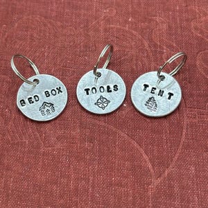 May include: Three circular silver metal tags, each attached to a small split ring. The tags are stamped with black text and symbols: one reads BED BOX with a mountain icon, one reads TOOLS with a compass icon, and one reads TENT with a tree icon.