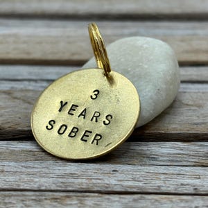 Può includere: Un'etichetta rotonda in metallo color oro con le parole "3 YEARS SOBER" stampate in nero. L'etichetta è attaccata a un anello color oro. L'etichetta poggia su una pietra bianca, su uno sfondo di legno.