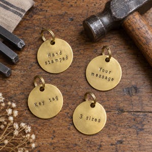 Könnte beinhalten: Vier kreisförmige Schlüsselanhänger aus Messing mit Metallringen, jeweils mit handgestempelter schwarzer Schrift. Die Anhänger zeigen die Texte Hand stamped, Your message, Key tag und 3 sizes. Ein Hammer und Stahlstempel sind ebenfalls zu sehen.