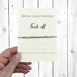 Puede incluir: Una tarjeta color crema con el texto "Morse code message" y "Fuck off" en fuente manuscrita. Una pulsera de código Morse con cuentas doradas y verdes está adjunta. La tarjeta también dice "Morse code bracelet with a hidden message" en la parte inferior.