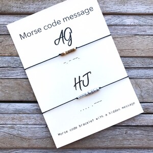 May include: Two friendship bracelets with a white card that says "Morse code message" and the initials "AG" and "HJ" written in cursive. The bracelets are made with black string and beads. The card also includes the Morse code for each initial.
