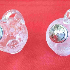 May include: Two clear glass salt shakers with etched floral designs and silver tops. The shakers have a round body and a handle.