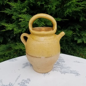 Peut inclure: Une théière en céramique bicolore jaune et beige avec un corps arrondi, une anse en boucle et un petit bec verseur. La théière a un couvercle et une petite poignée sur le côté. Le fond est un buisson vert.