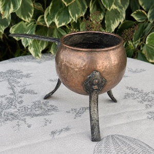 May include: A hammered copper pot with a rounded body and a dark, textured metal handle. The pot is supported by three dark, riveted metal legs that extend from the base. The interior of the copper vessel is dark, and the exterior surface shows a distinct, hand-hammered texture throughout the metal.