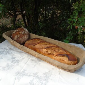 Puede incluir: Un cuenco de madera rústico contiene dos panes sobre un mantel blanco. Uno es redondo y el otro es una baguette larga. La escena está ambientada al aire libre, con vegetación de fondo. La baguette mide unos 40 cm.