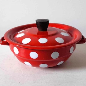 Puede incluir: Una cacerola de esmalte roja con tapa, adornada con lunares blancos. El plato tiene dos asas pequeñas y un pomo negro en la tapa. Un artículo de cocina clásico.