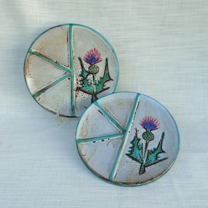 Può includere: Due piatti rotondi in ceramica con sfondo grigio chiaro e dettagli turchese. Ogni piatto presenta un motivo di cardo con dettagli viola e verdi. I piatti sono divisi da linee turchesi.