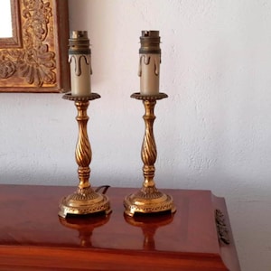 Może przedstawiać: Para złotych metalowych podstaw lamp w kształcie świecznika z białymi kloszami. Podstawy mają skręcony wzór i okrągłą podstawę.
