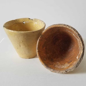 May include: Two antique, handmade, yellow-toned ceramic pots. One is upright with a hole near the rim, the other is lying on its side. The interior of the pots is a dark brown colour, and the rims have a textured, aged appearance.