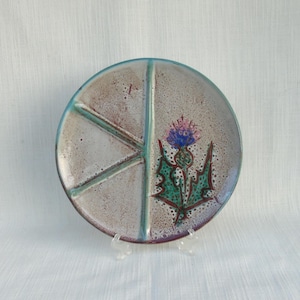 May include: A round, divided plate with a hand-painted thistle design. The plate is divided into four sections by turquoise lines. The thistle features a purple and blue flower with green leaves. The plate has a speckled, off-white background and a turquoise rim.