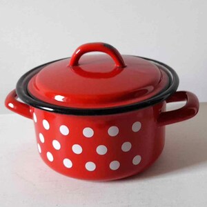 Może przedstawiać: Czerwony emaliowany garnek z pokrywką i dwoma bocznymi uchwytami. Garnek jest ozdobiony białymi kropkami. Obręcz i uchwyty są czarne. Garnek stoi na białej powierzchni.