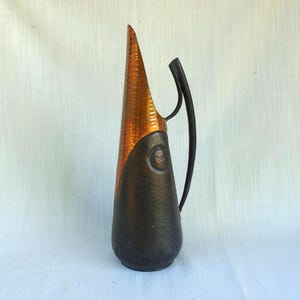Puede incluir: Jarra decorativa con parte superior de color cobre y base marrón oscuro. La sección de cobre tiene un aspecto texturizado y martillado. Un asa negra se curva desde la parte superior hacia el lado. La jarra tiene una forma única y alargada.