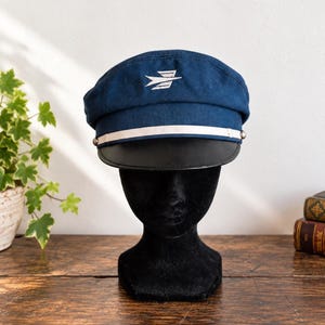 Könnte beinhalten: Eine marineblaue Schirmmütze mit einem weißen, gestickten Flügel-Emblem auf der Vorderseite. Die Mütze verfügt über ein weißes horizontales Band an der Basis der Krone, das mit silberfarbenen Knöpfen an den Seiten befestigt ist, und einen schwarzen Schirm.