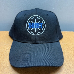 Pode incluir: Boné de beisebol preto com aba curva. O boné apresenta um emblema circular com o texto "501st" em azul, rodeado por um design branco. O boné é feito de um tecido texturizado.