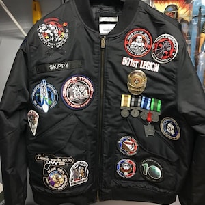 May include: A black bomber jacket with a variety of embroidered patches, including Star Wars themed patches, a military patch, and a patch that says "SKIPPY".