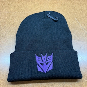 May include: Black knit beanie with a purple embroidered emblem. The beanie has a folded brim and a small black tag attached. The emblem is a stylized, geometric design.