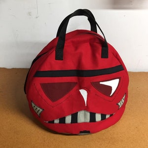 以下が含まれることがあります： 黒のハンドルとストームトルーパーのヘルメットの白黒のデザインが施された赤いキャンバスバッグ
