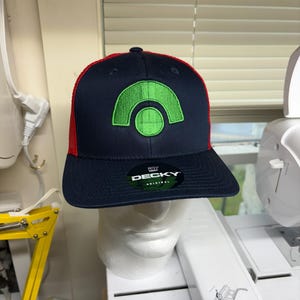 Könnte beinhalten: Eine marineblaue Baseballkappe mit rotem Netzrücken und einem leuchtend grünen, gestickten bogenförmigen Logo auf der Vorderseite. Die Kappe hat einen flachen Schirm mit einem schwarzen, runden Aufkleber, der den weißen Text DECKY ORIGINAL trägt.