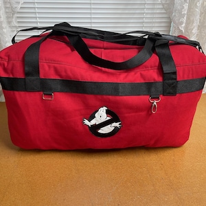 Może przedstawiać: Czerwona torba podróżna z czarnymi paskami i akcentami. Torba posiada logo Ghostbusters z przodu. Torba ma prostokątny kształt i jest przeznaczona do przenoszenia przedmiotów. Torba stoi na brązowej powierzchni.