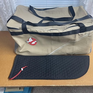 Może przedstawiać: Torba podróżna z płótna w kolorze beżowym z czarnymi paskami i logo Ghostbusters. Torba ma czarną, pikowaną kieszeń zapinaną na zamek błyskawiczny.