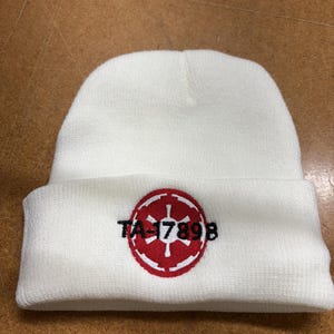 Pode incluir: Um gorro branco de malha com um emblema circular vermelho e branco e o texto "TA-1789B". O emblema é um design estilizado. O gorro é feito de um material macio e texturizado.