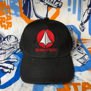 Peut inclure: Casquette de baseball noire avec un logo rouge et blanc et le mot "ROBOTECH" brodé en rouge. La casquette a une visière incurvée et est faite d'un tissu durable. C'est un article de fan.