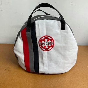 May include: White canvas bag with black handles and red, gray, and black stripes. The bag has an embroidered red and white logo with the text "DA12904".