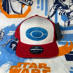 Könnte beinhalten: Rote, weiße und blaue Baseballkappe mit Mesh-Rückseite. Die Vorderseite der Kappe ist weiß mit einem blauen Oval-Design. Der Schirm ist rot und die Kappe hat ein "DECKY"-Etikett.