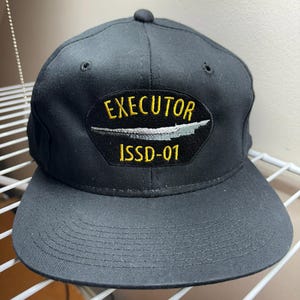 Puede incluir: Gorra de béisbol negra con visera curva. La gorra presenta un parche con la palabra "EXECUTOR" en letras amarillas sobre una imagen estilizada y el texto "ISSD-01" en amarillo.