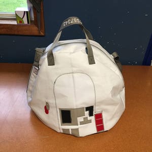 以下が含まれることがあります： グレーのハンドルとグレーのストラップが付いた白いキャンバスバッグ。バッグの前面には赤、グレー、黒のデザインが施されています。ハンドルには「1557343」という文字が刺繍されています。