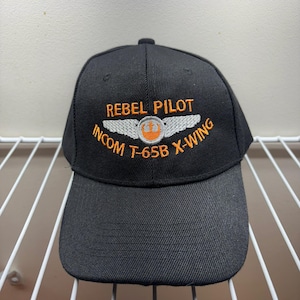 May include: Black baseball cap with the text "REBEL PILOT INCOM T-65B X-WING" embroidered in orange and white. The cap features a curved brim and a rounded top. The design includes a stylized emblem of a ship.