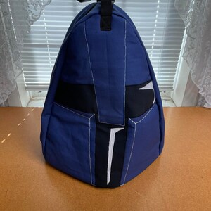 以下が含まれることがあります： 黒と白のアクセントが付いた青いキャンバスのバックパック。バックパックはヘルメットのようなユニークなデザインです。
