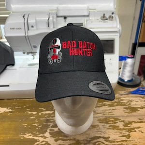 May include: Black baseball cap with the text "BAD BATCH HUNTER" embroidered in red. The cap features a graphic of a helmet. The hat is displayed on a mannequin head.