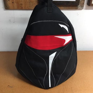 May include: Black backpack with a red and white design resembling a helmet. The design features a white stripe and a red band across the top.
