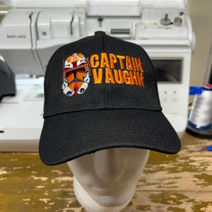 Op de afbeelding: Zwarte baseballcap met de tekst "CAPTAIN VAUGHN" in oranje en een gedetailleerd geborduurd ontwerp van een helm. De cap wordt getoond op een witte mannequin hoofd.