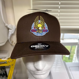 May include: A brown baseball cap with a structured crown and a flat brim. The front features an embroidered patch with a golden eagle, red, white, and blue accents, and the text FALL BUY STUNTMAN ASSOC. A black DECKY ORIGINAL sticker is on the brim.