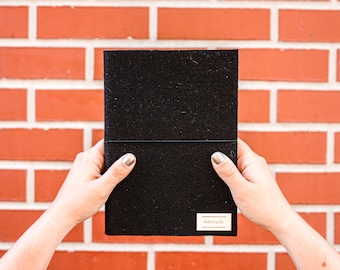 May include: A black felt notebook with a gold label that says "kerouch" on a brick wall background.