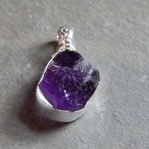 May include: A silver pendant featuring a raw amethyst gemstone. The amethyst is a deep purple color and has a natural, rough texture.