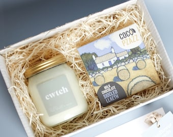 Könnte beinhalten: Eine weiße Geschenkbox gefüllt mit zerkleinertem Papier, eine weiße Kerze in einem Glasgefäß mit goldenem Deckel und einem Etikett mit der Aufschrift "ewtch" sowie eine Tafel Schokolade mit einer bunten Illustration eines Hauses und Heuballen. Die Schokoladentafel ist mit "Coco Piazz" und "Milchschokolade Siocled Llaeth" beschriftet.