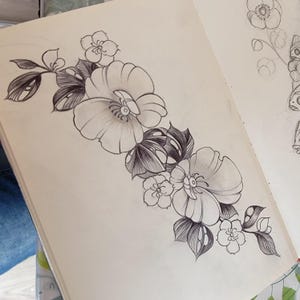 Op de afbeelding: Een gedetailleerde pen- en inkttekening van een bloemenontwerp. De illustratie toont grote, open bloemen met gedetailleerde bloemblaadjes en kleinere bloesems, samen met bladeren. Het kunstwerk staat op crèmekleurig papier, met andere schetsen zichtbaar op de achtergrond.
