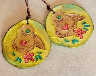 May include: Two circular pendants with a bird design. The birds are brown with a red heart and green eye. Pink flowers and green leaves surround the bird. Each pendant is attached to a brown string.