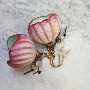 May include: A pair of handmade earrings featuring a unique floral design. The earrings showcase pink and white petal-shaped elements with a light green interior. They are accented with gold-colored metal and small, clear beads.