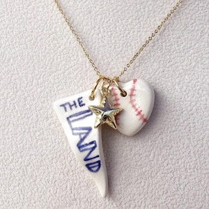 May include: A gold chain necklace featuring three charms: a white triangular pendant with the blue text THE LAND, a small gold star, and a white heart-shaped pendant with red baseball-style stitching details.