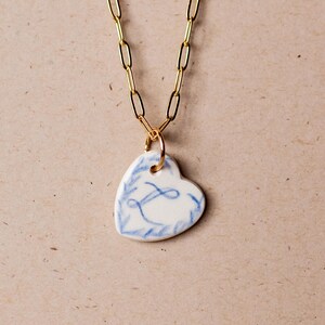 May include: A gold chain necklace with a white porcelain heart pendant. The heart has a blue floral design and the letter "L" in the center.