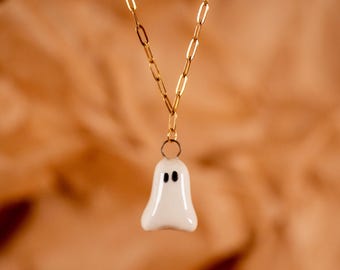 May include: A gold-colored chain necklace with a white ghost pendant. The ghost pendant has two black dot eyes. The background is a blurred orange-brown color.