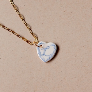 May include: A gold chain necklace with a white ceramic heart pendant. The heart has a blue floral design and the letter "L" in the center.